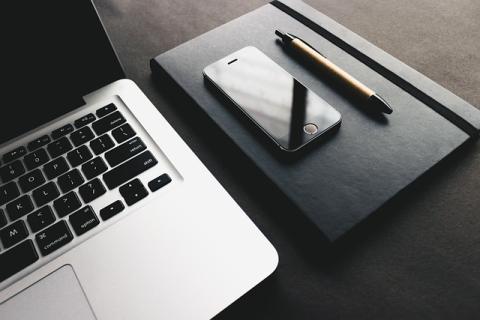 Overhead partial view of a laptop screen/keyboard, smart phone laying next to a pen