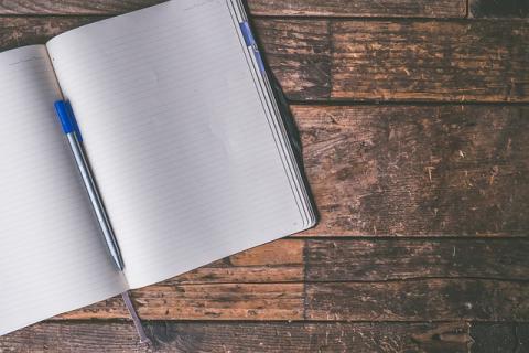 An open notebook with lined paper on a wooden table. A pen rests vertically in the spine of the notebook.