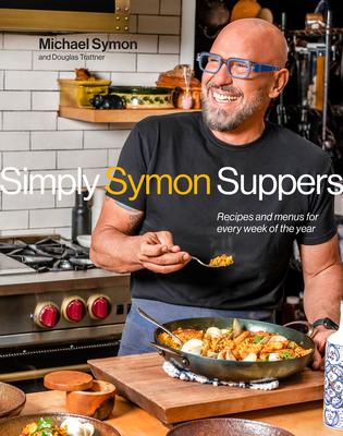 Smiling bald man with a grey beard and blue-framed glasses holding a forkful of food in his right hand, standing over a pan of food on top of a cutting board on a kitchen counter