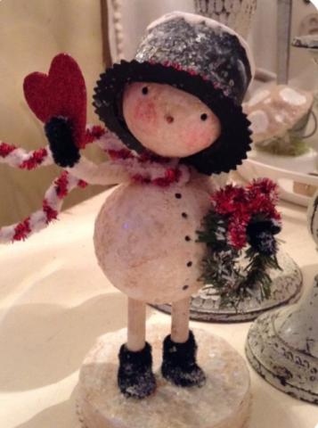 Little snowman with legs and arms, wearing a hat and holding holly in one hand and a large heart in the other