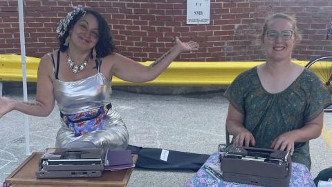 Two women sitting at a table with typewriters in front of them, the one on the right wearing a silver dress with her hands in the air, the one on the right smiling and typing, wearing a brown dress