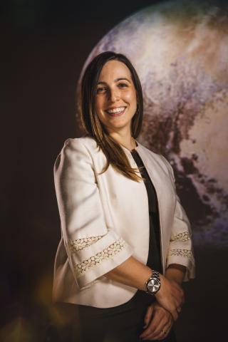 Waist up portrait of woman in a white coat with brown hair, smiling, her right hand clasping her left wrist