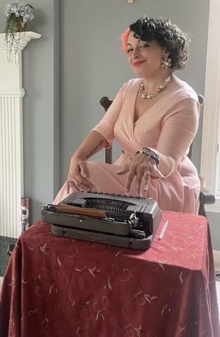 Woman in a pink dress sitting at a table with a vintage typewriter.