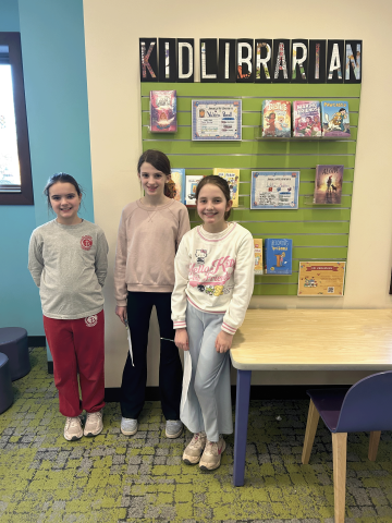 Three kids stand in front of the Kid Librarian display at the library