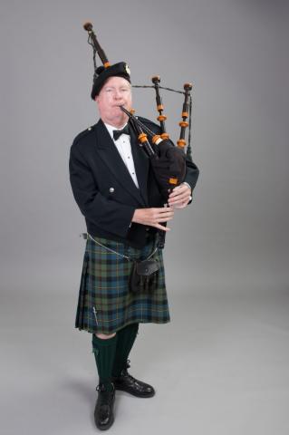 Man holding bagpipes, dressed in full regalia, a beret, suit coat, kilt, high socks and black shoes.