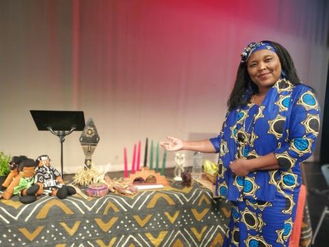A woman in a colorful dress stands beside a table full of traditional Kwanzaa items, such as dolls, candles etc.