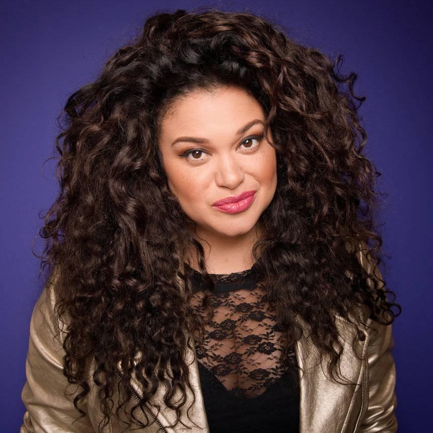 Portrait of a smiling woman with long, wavy hair, wearing a gold jacket 