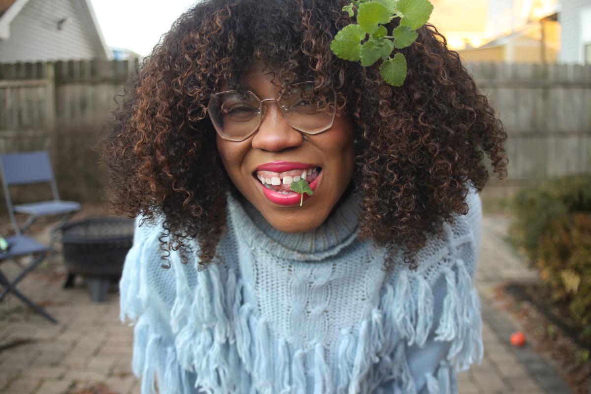 A portrait of a woman smiling, with a foraged plant beneath her teeth and leaves in her hair